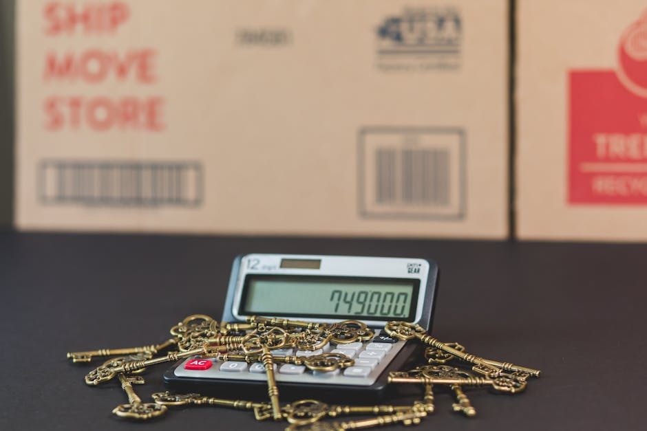 A digital calculator displaying the number 749800 is placed on a dark surface, surrounded by a scattered collection of antique brass keys and a few keychains. Behind the calculator, there are several