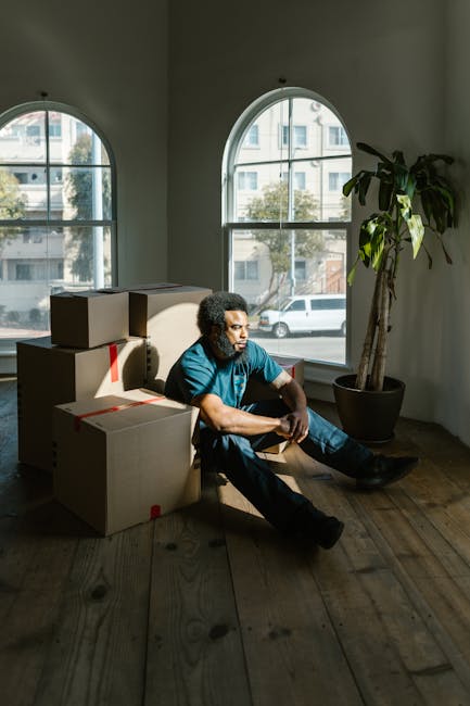A man with curly hair and a beard, dressed in a blue t-shirt and dark trousers, sits on a wooden floor inside a room with large arched windows revealing a street view outside. He is surrounded by mult
